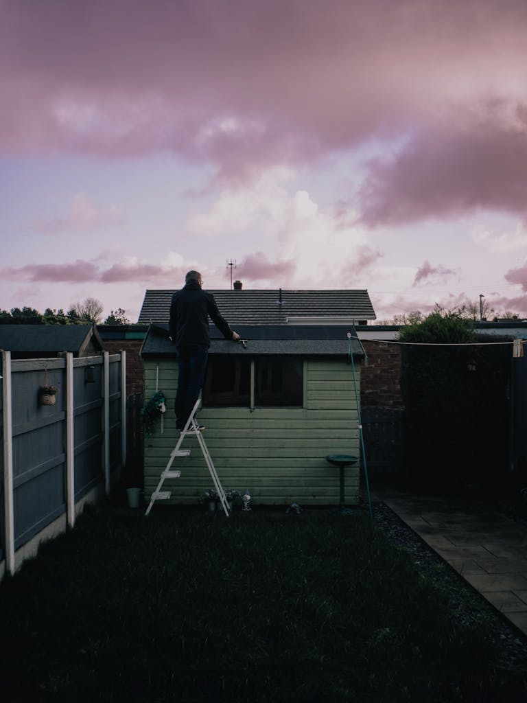 A man fixing the roof of a small shed in a backyard at dusk with a dramatic sky.