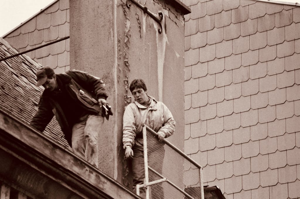 Two construction workers on a rooftop performing repair work, captured in sepia tones.